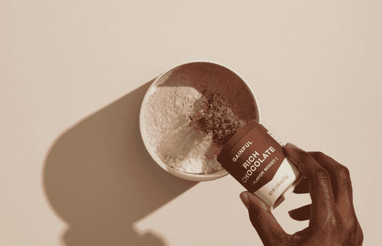 A hand mixing a small jar of flavor powder with a larger jar of unflavored protein